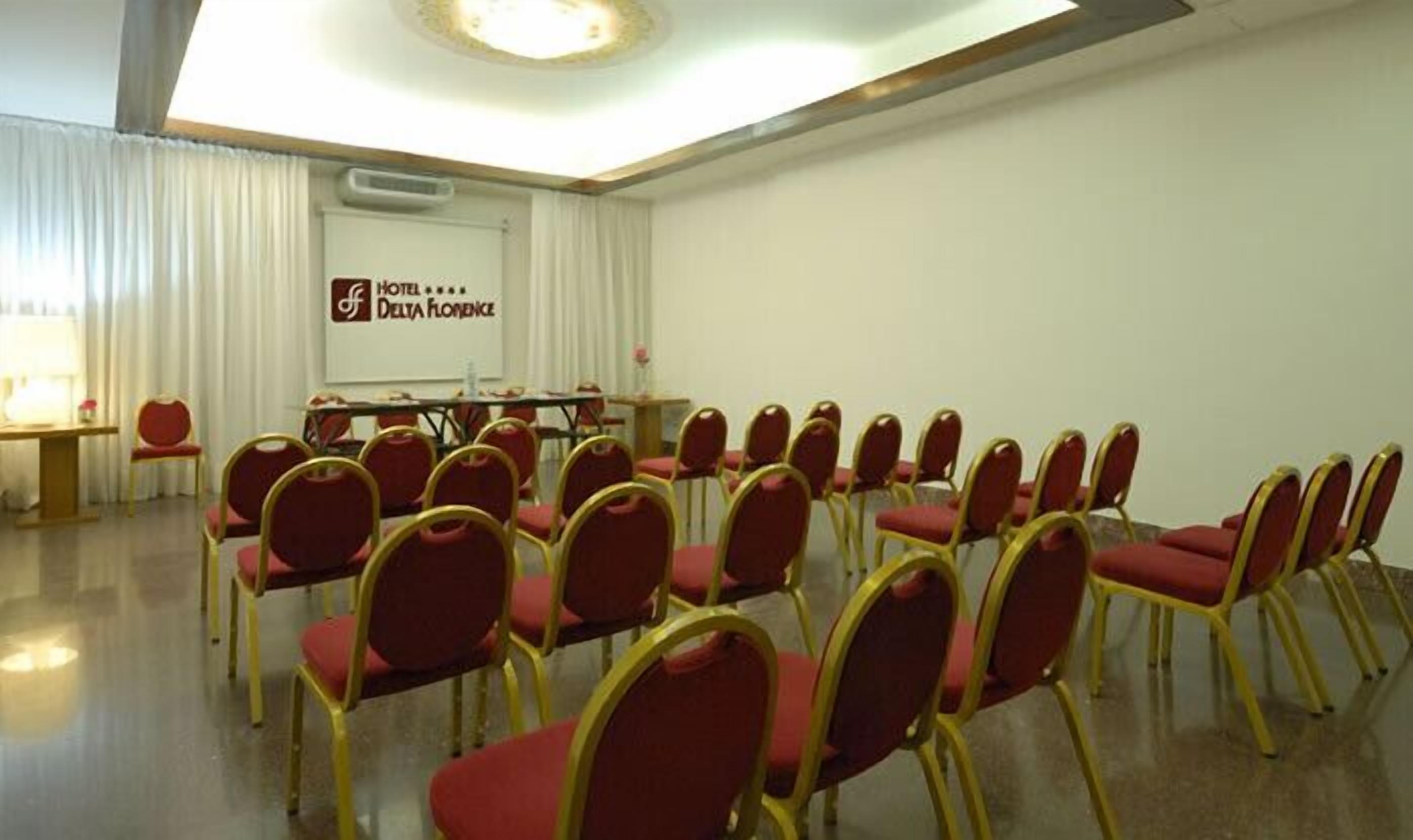 A conference room with rows of red chairs and a table, a whiteboard, and a sign for Hotel Delta Florence.