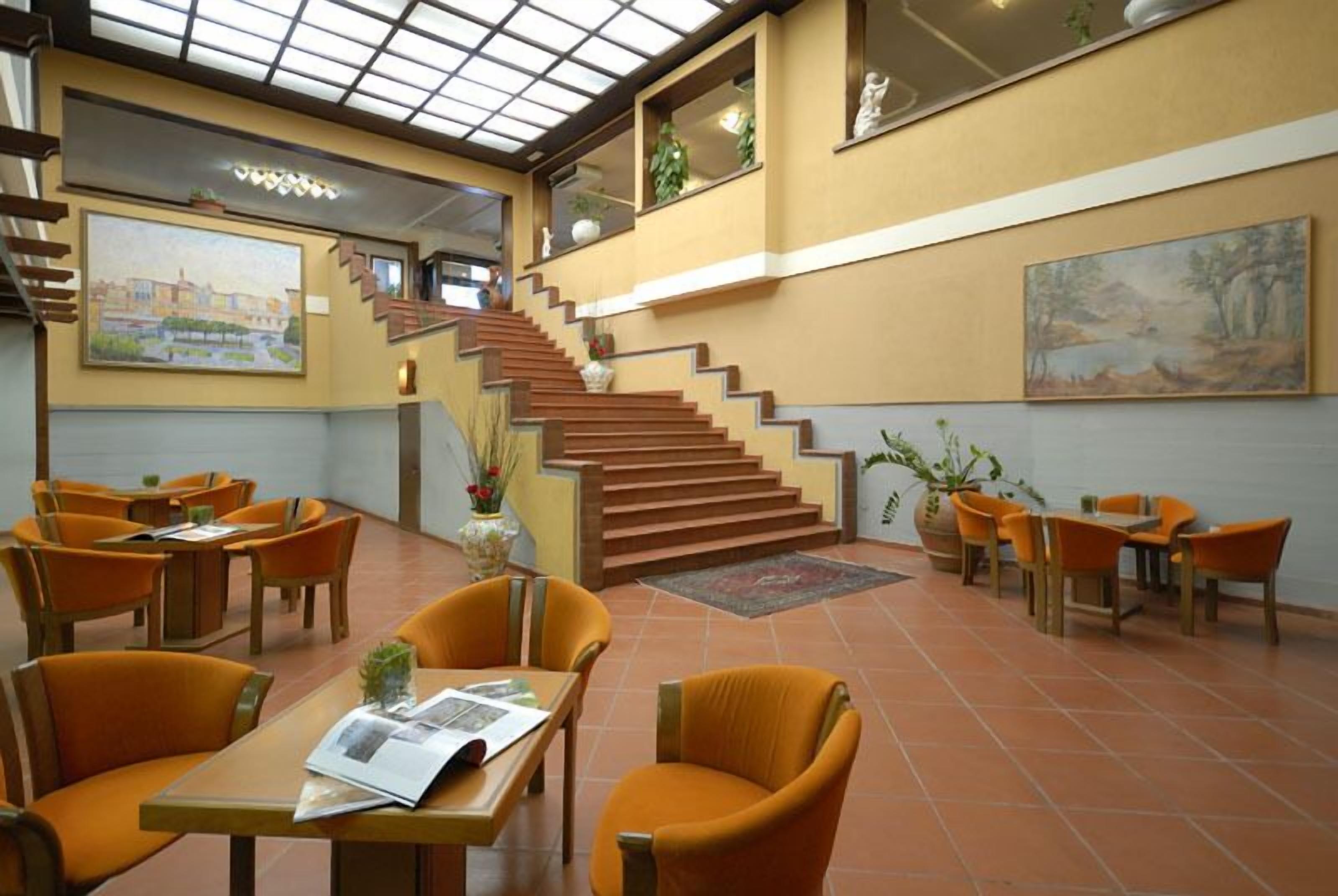 A hotel lobby with a staircase, orange chairs, and tables.