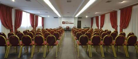 A conference room with rows of chairs, a podium, and a banner reading "Hotel de Draga Resort".