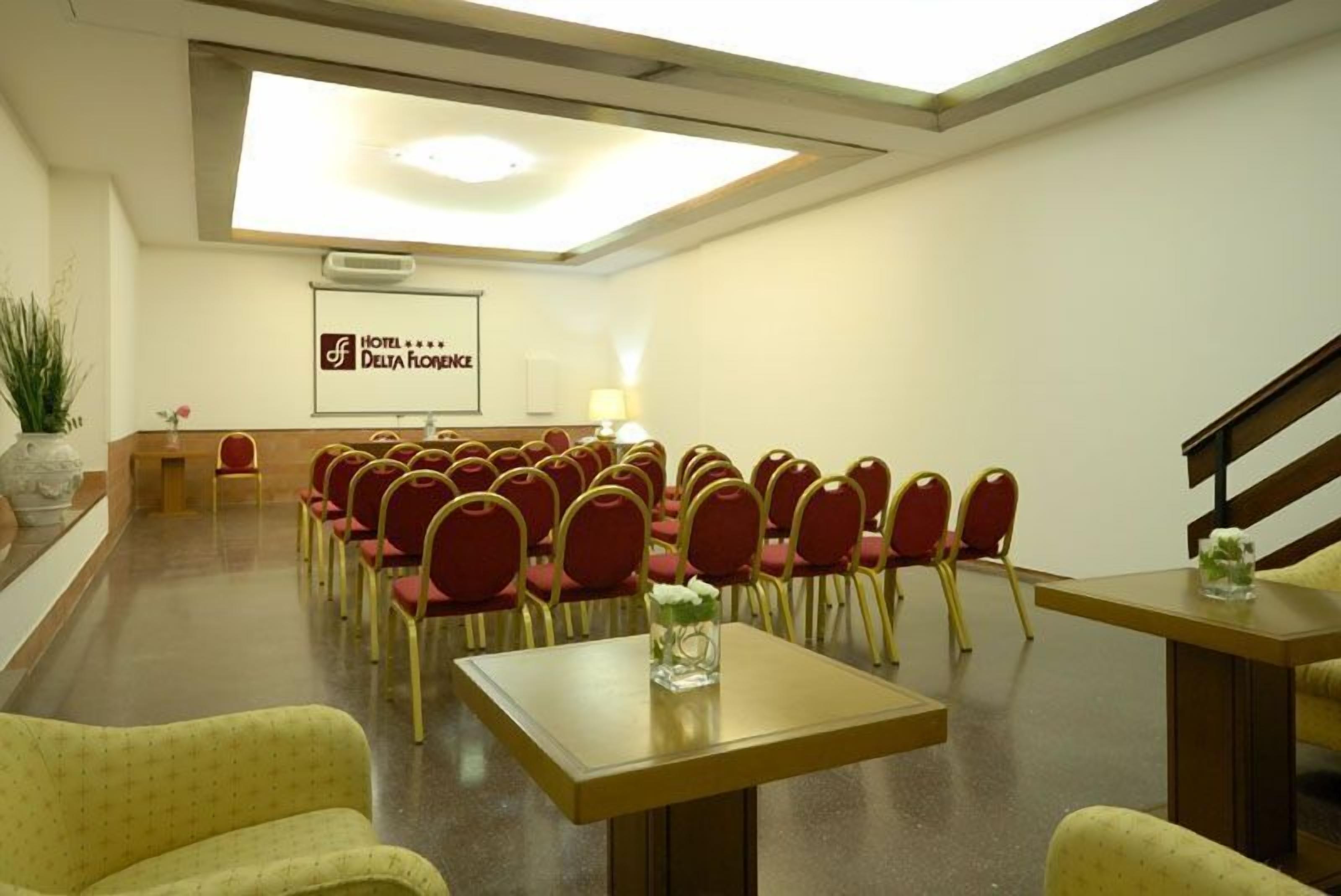 Un salón de conferencias de hotel con filas de sillas rojas, una mesa de madera y una pantalla proyectando el nombre del hotel.