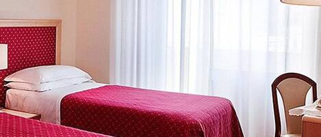 A hotel room with two beds, a wooden nightstand, a chair, and a window with sheer curtains.