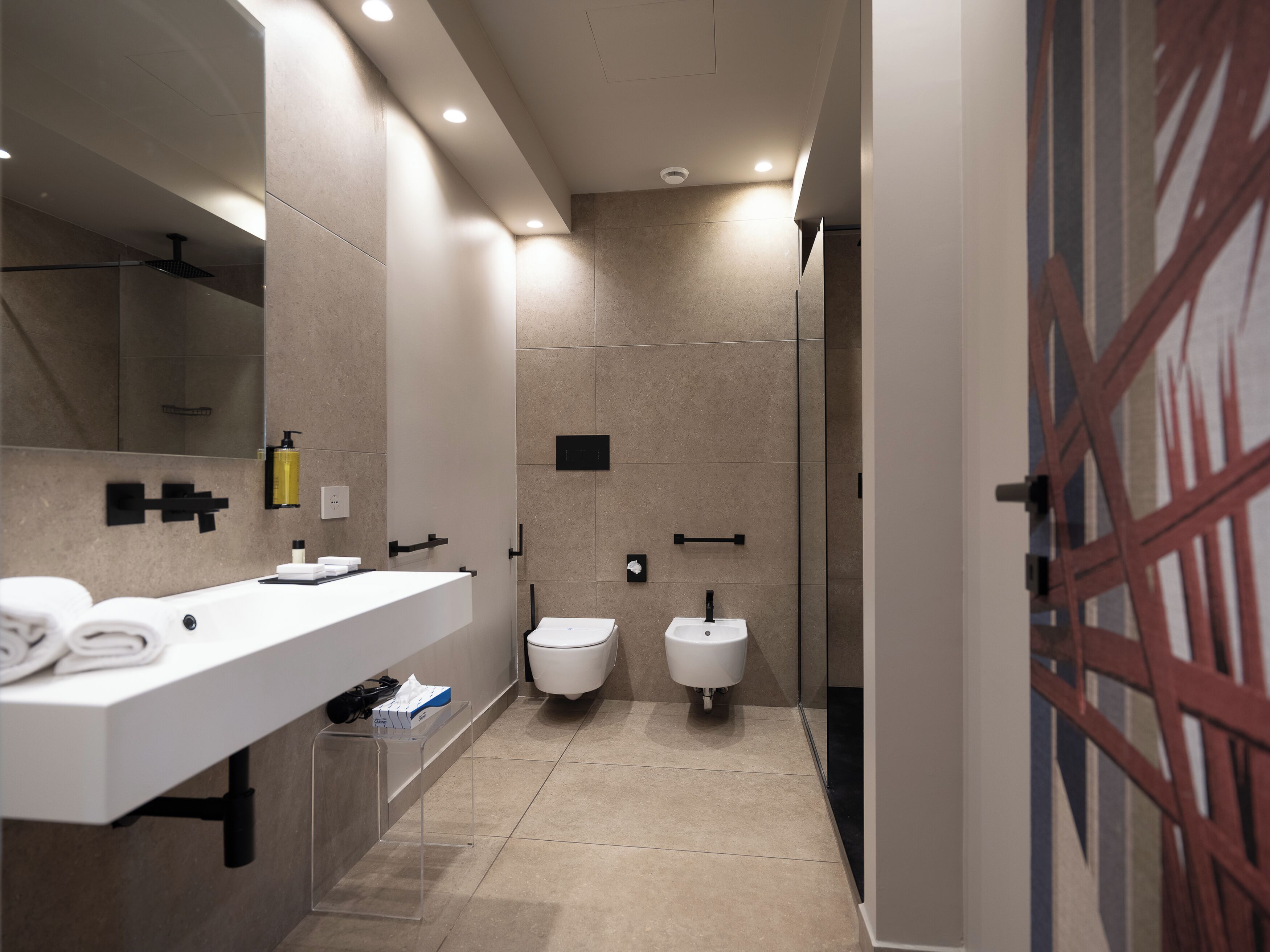 Une salle de bain moderne avec un lavabo blanc, deux toilettes, une douche et une porte en verre.