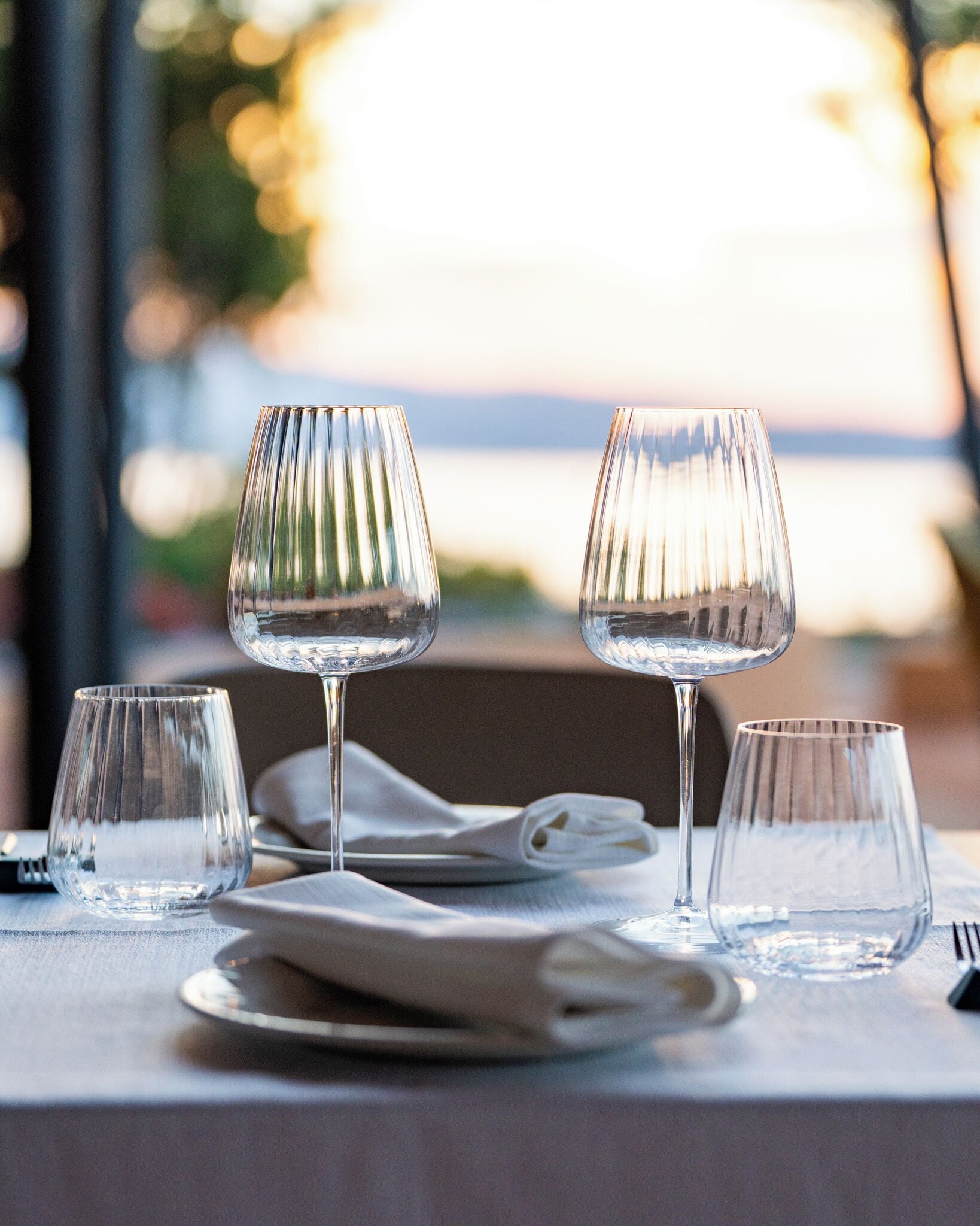 Una mesa servida con cuatro copas de vino y platos, con vista al agua de fondo.