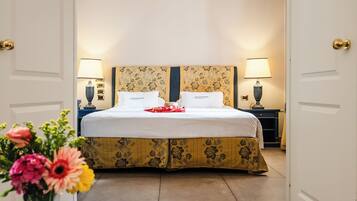 Habitación de hotel con cama, lámparas de noche, un florero con flores y un libro sobre una mesa de vidrio.