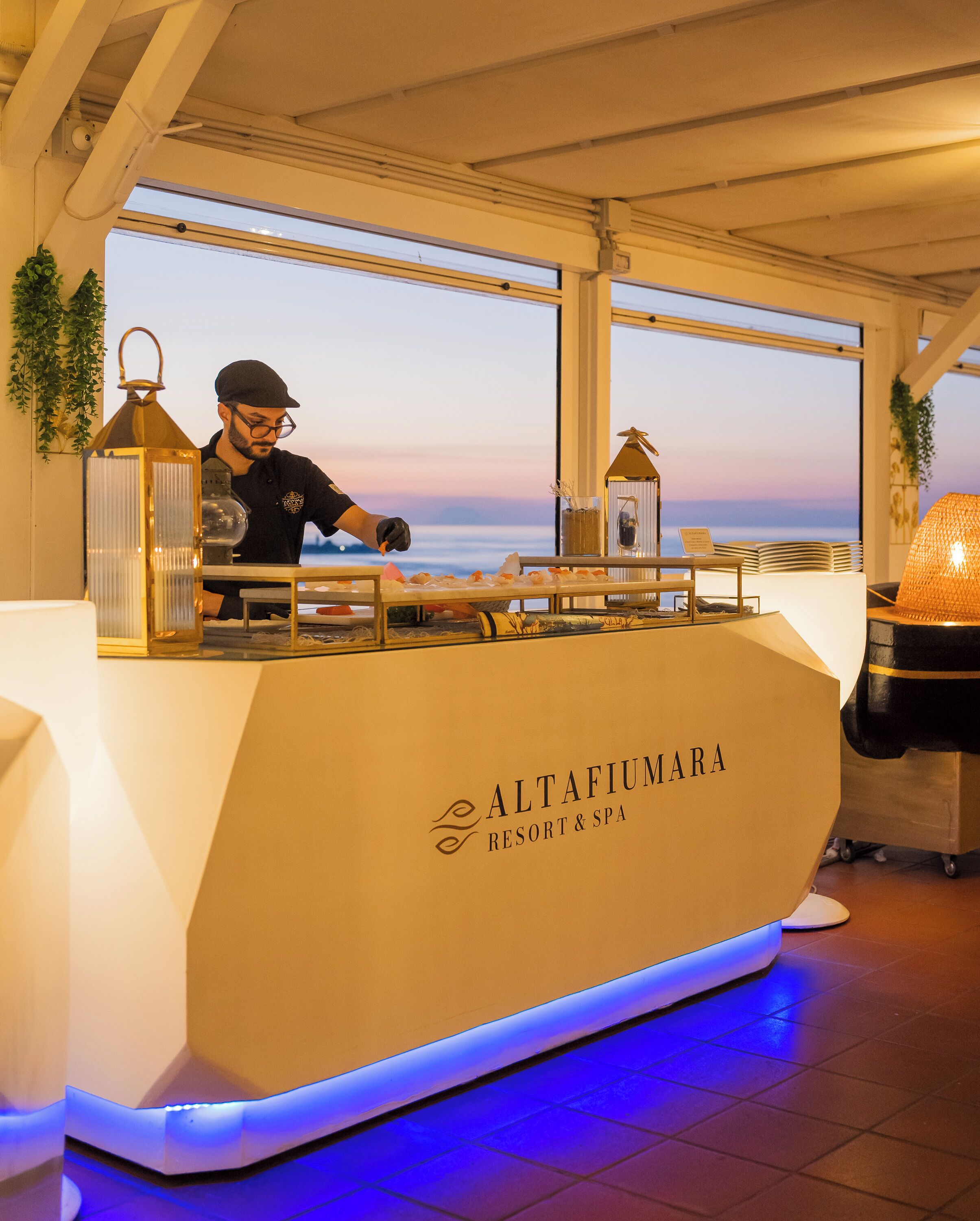 A bar area with a view of the sea, a sign reading 'Altafiumara Resort & Spa', and a person preparing food.