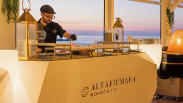 A bar area with a view of the sea, a sign reading 'Altafiumara Resort & Spa', and a person preparing food.