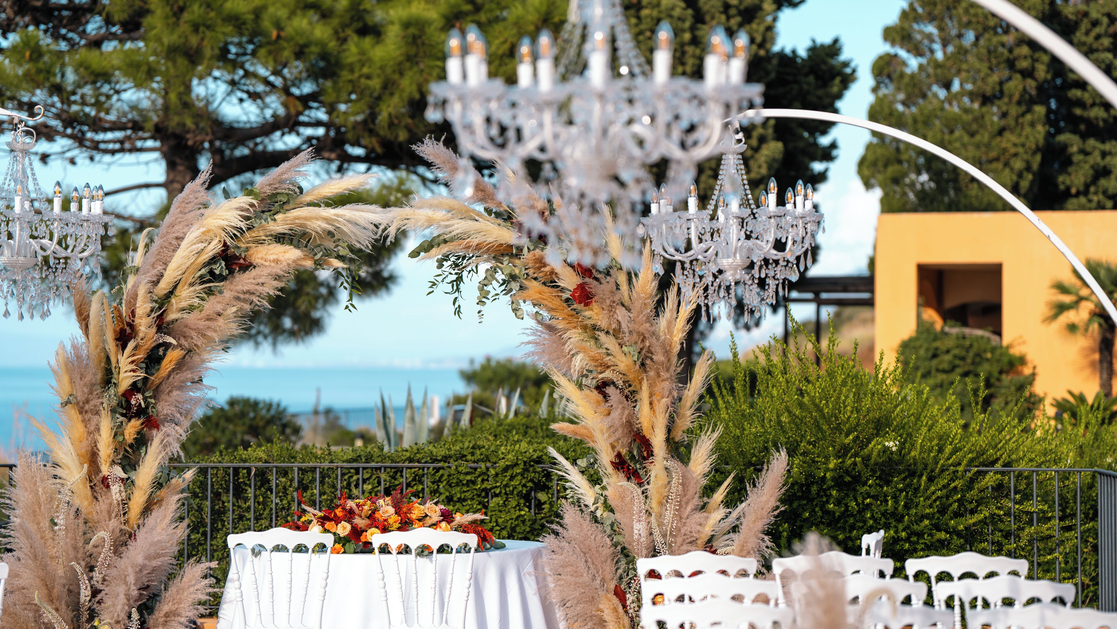 An outdoor wedding setup with a white table, chairs, and a decorative arch of pampas grass.