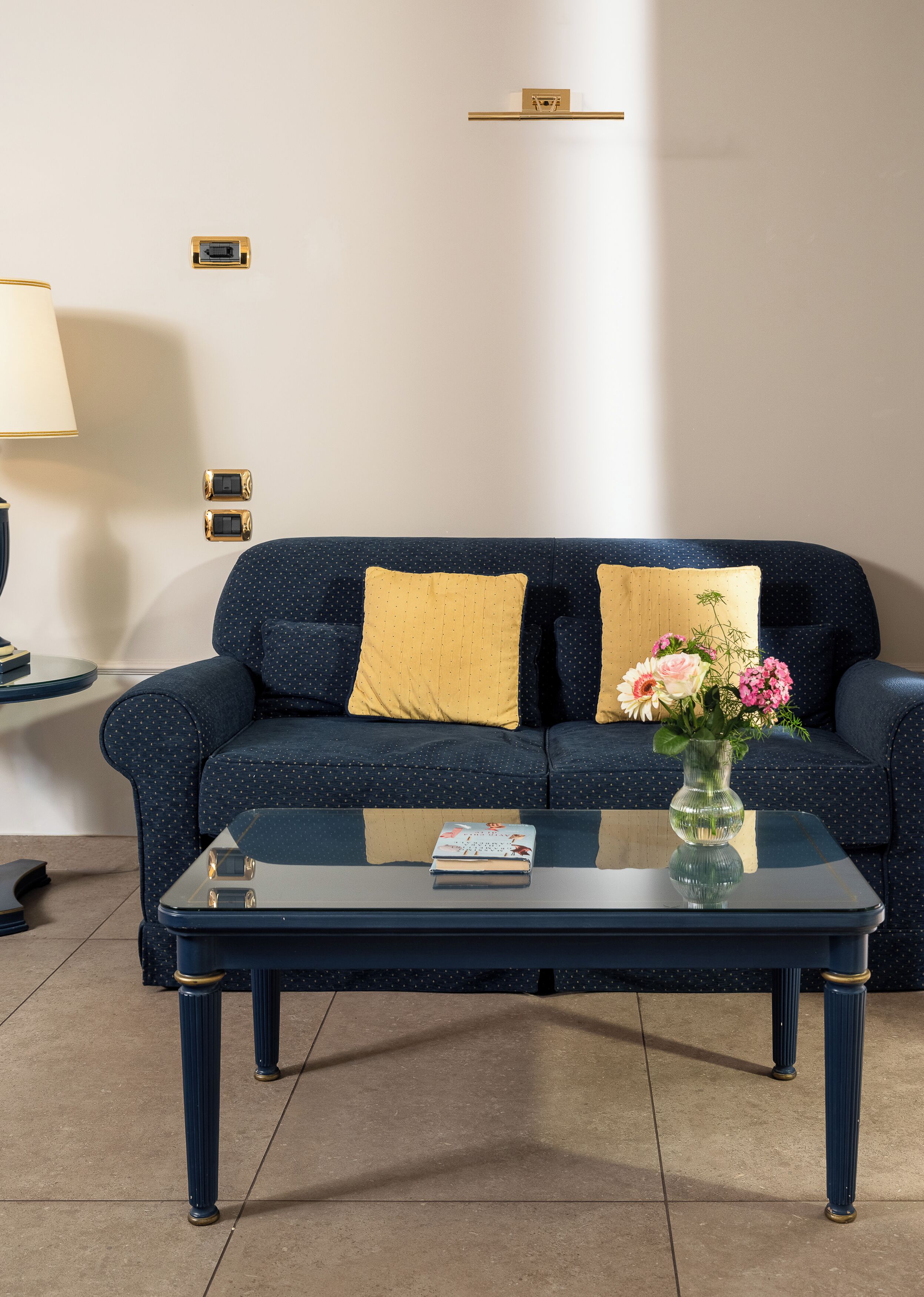 A dark blue sofa with yellow and white pillows, a glass coffee table with a book and a vase of flowers, and a lamp on a side table.