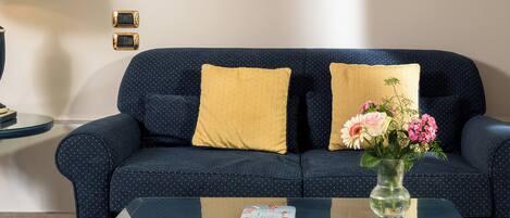 A dark blue sofa with yellow and white pillows, a glass coffee table with a book and a vase of flowers, and a lamp on a side table.