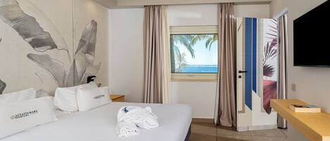 Habitación de hotel con cama, almohadas, toalla, televisor, ventana con cortinas y vista al mar.