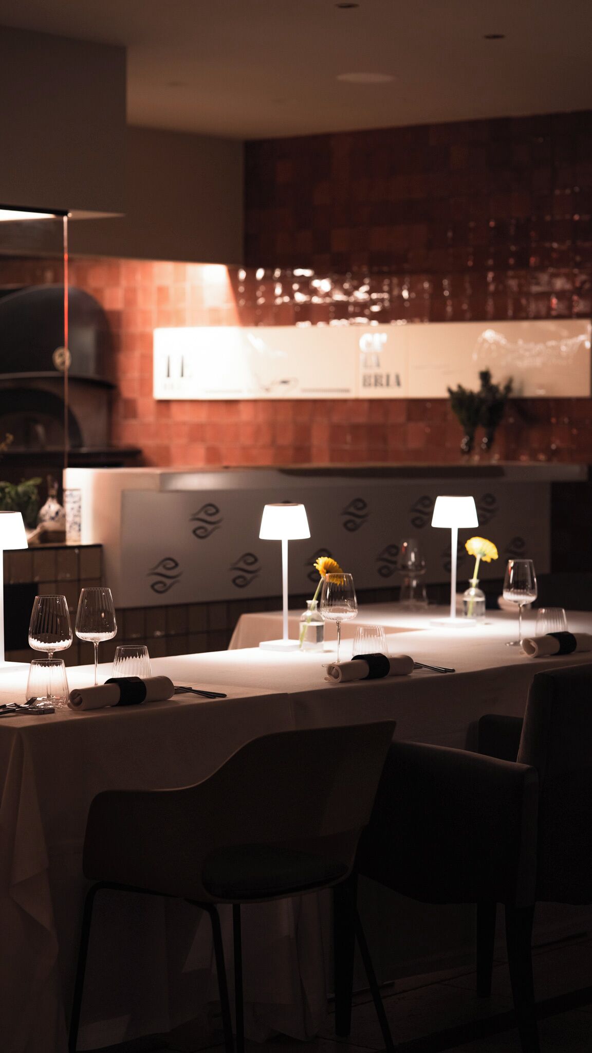 Ein schummrig beleuchtetes Restaurant mit einem langen Esstisch, das ein modernes Design mit einer Ziegelwand und einer großen Speisekarte im Hintergrund aufweist.