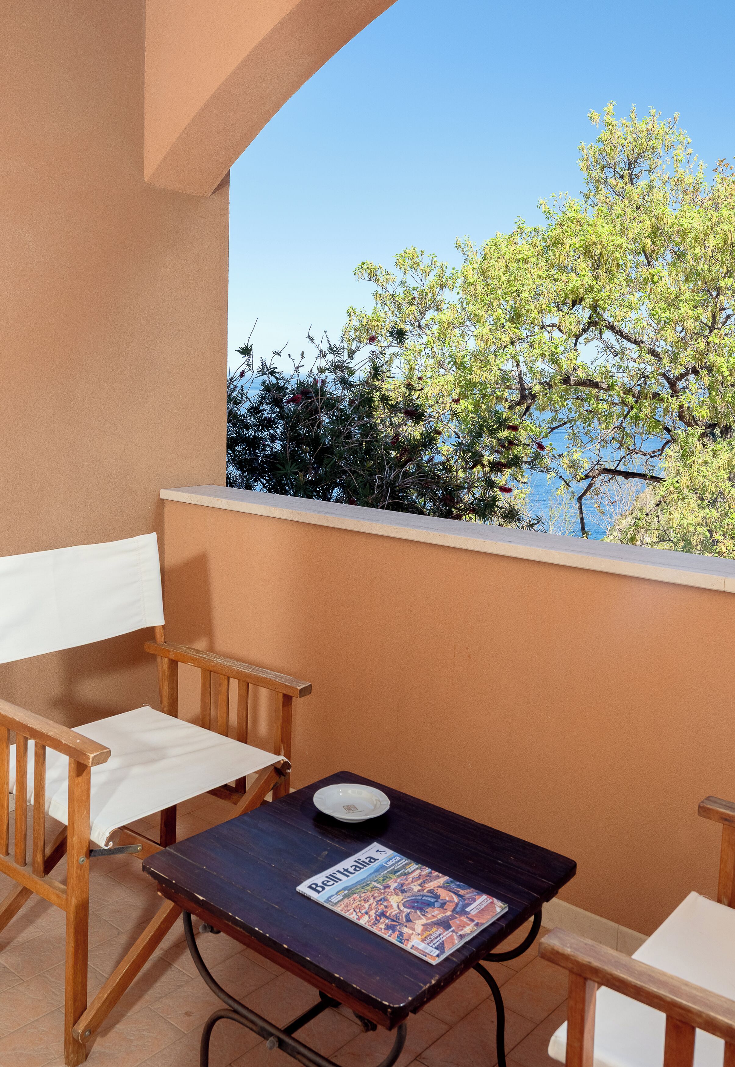 A balcony with a wooden chair, a small table with a magazine, and a view of trees and a body of water.