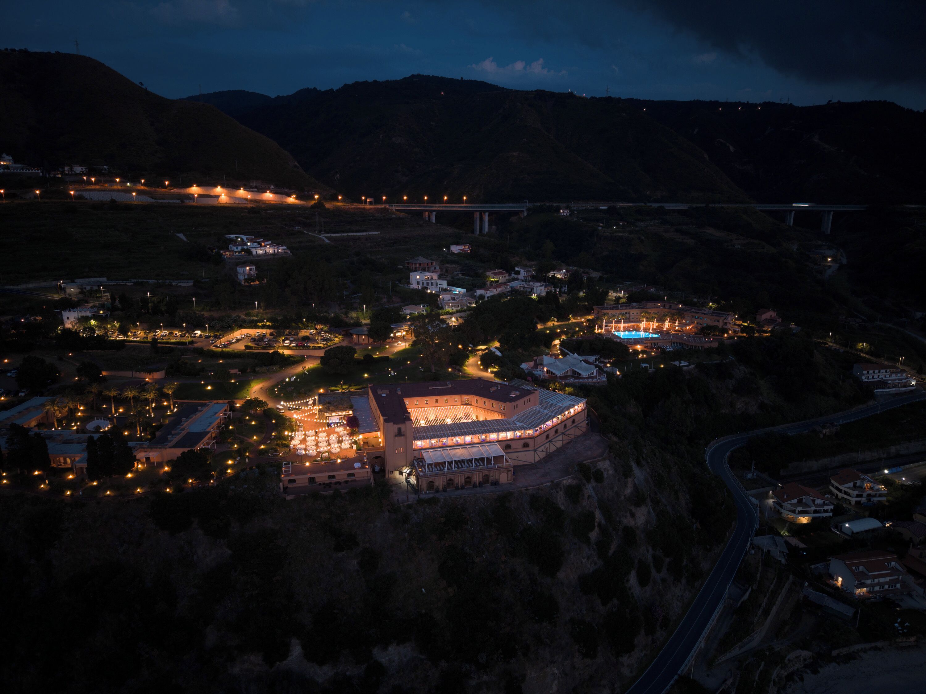An aerial view of a town at night with a prominent building with a large roof and multiple windows, surrounded by smaller buildings and illuminated streets.