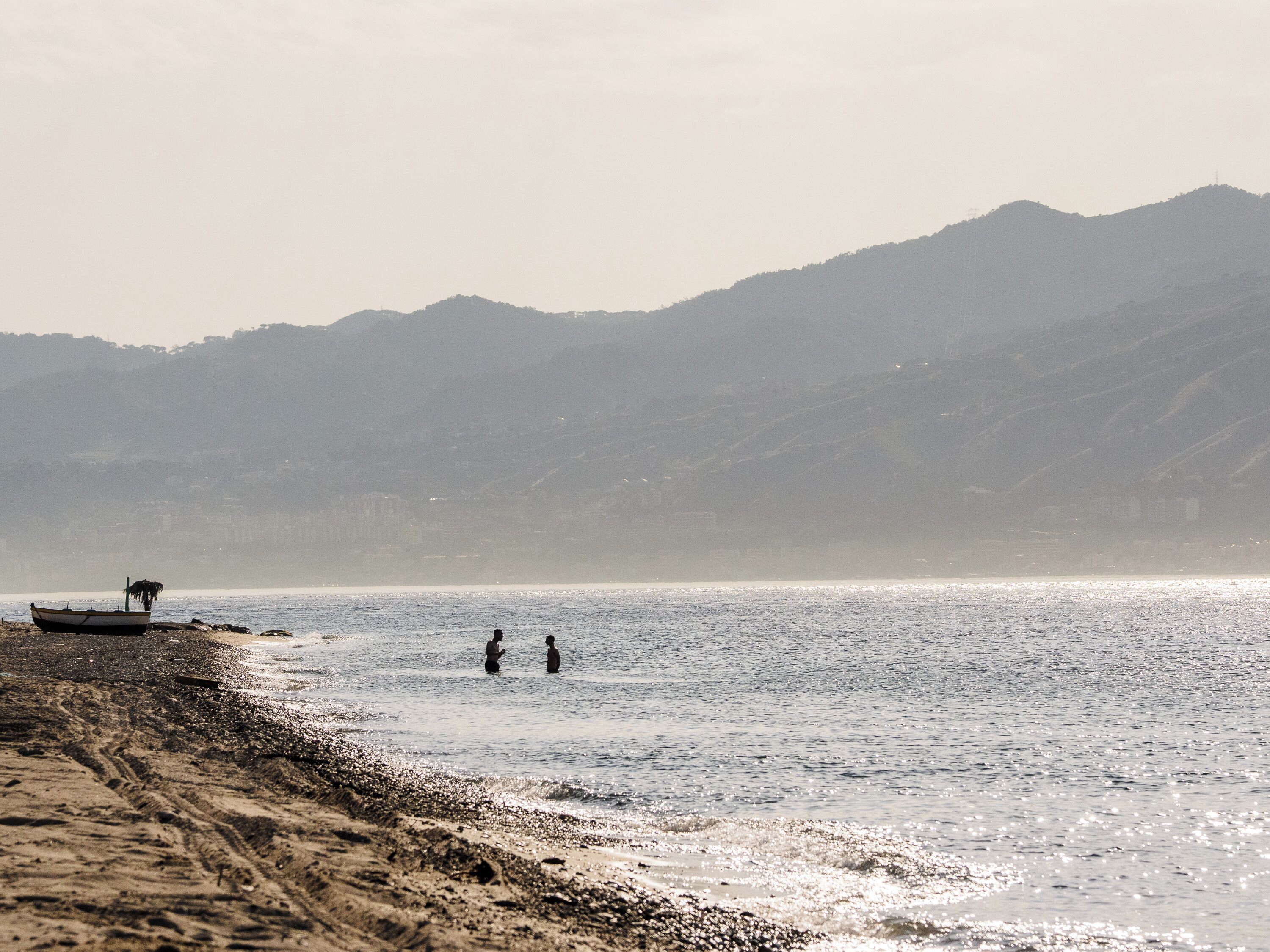 Una playa con un barco en la orilla, una persona de pie en el agua y montañas al fondo.