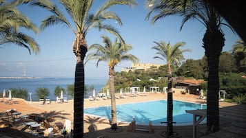 Zona junto a la piscina con sillones de descanso y sombrillas, palmeras y vista al mar.