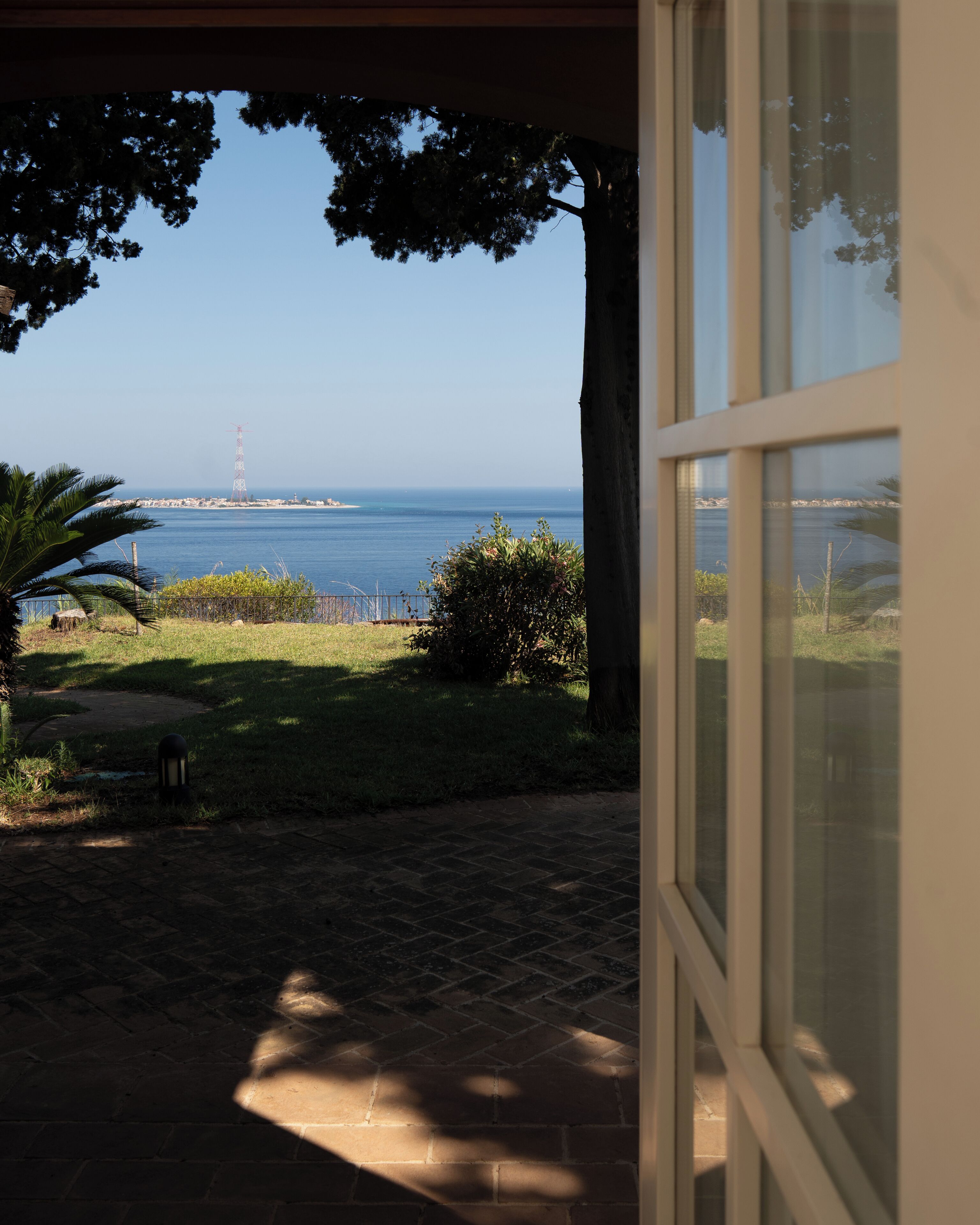 A view from a window overlooking a grassy area with a paved path, a palm tree, and a body of water in the distance.