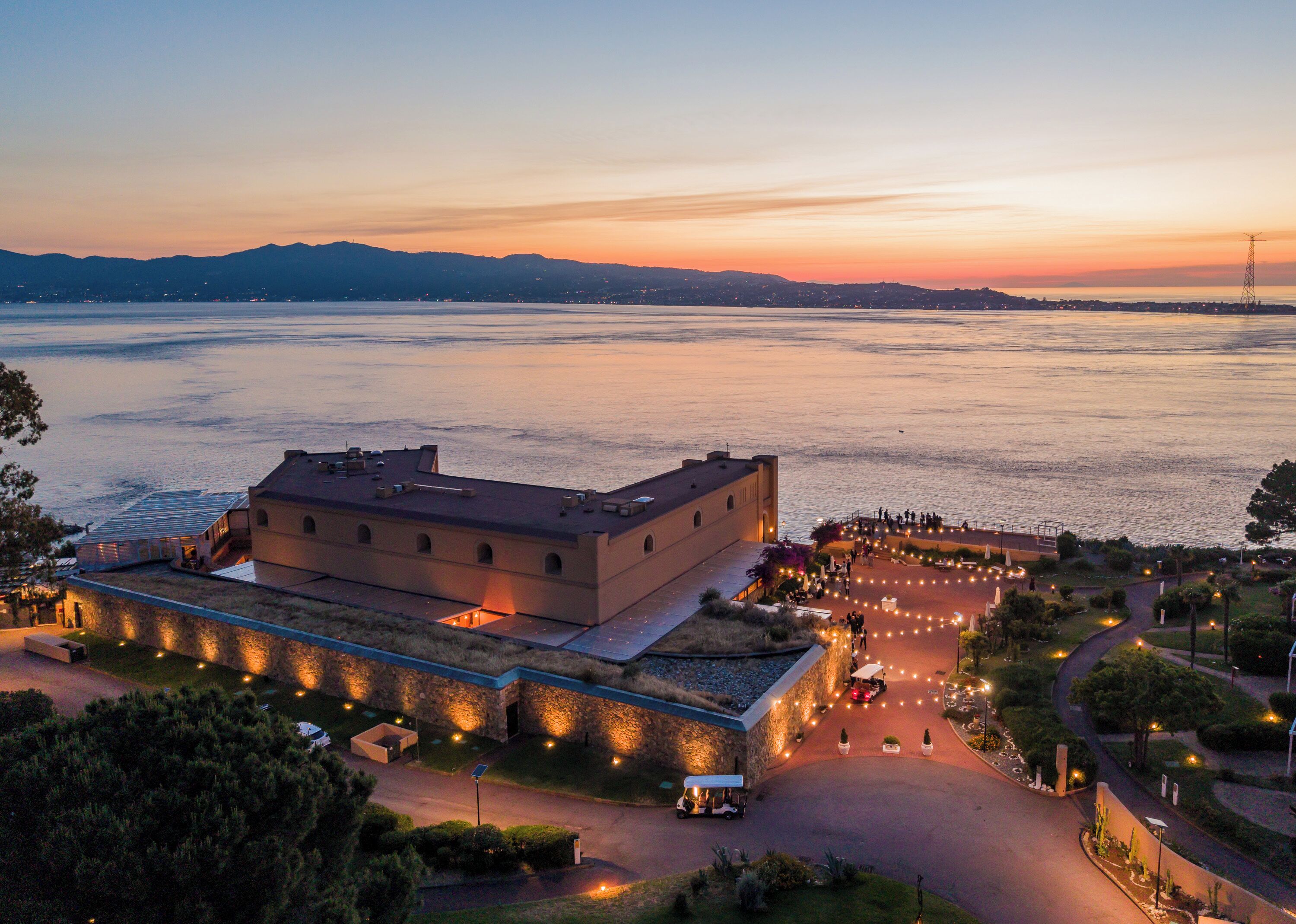 A large building with a flat roof, a paved area with lights, and a view of the water.