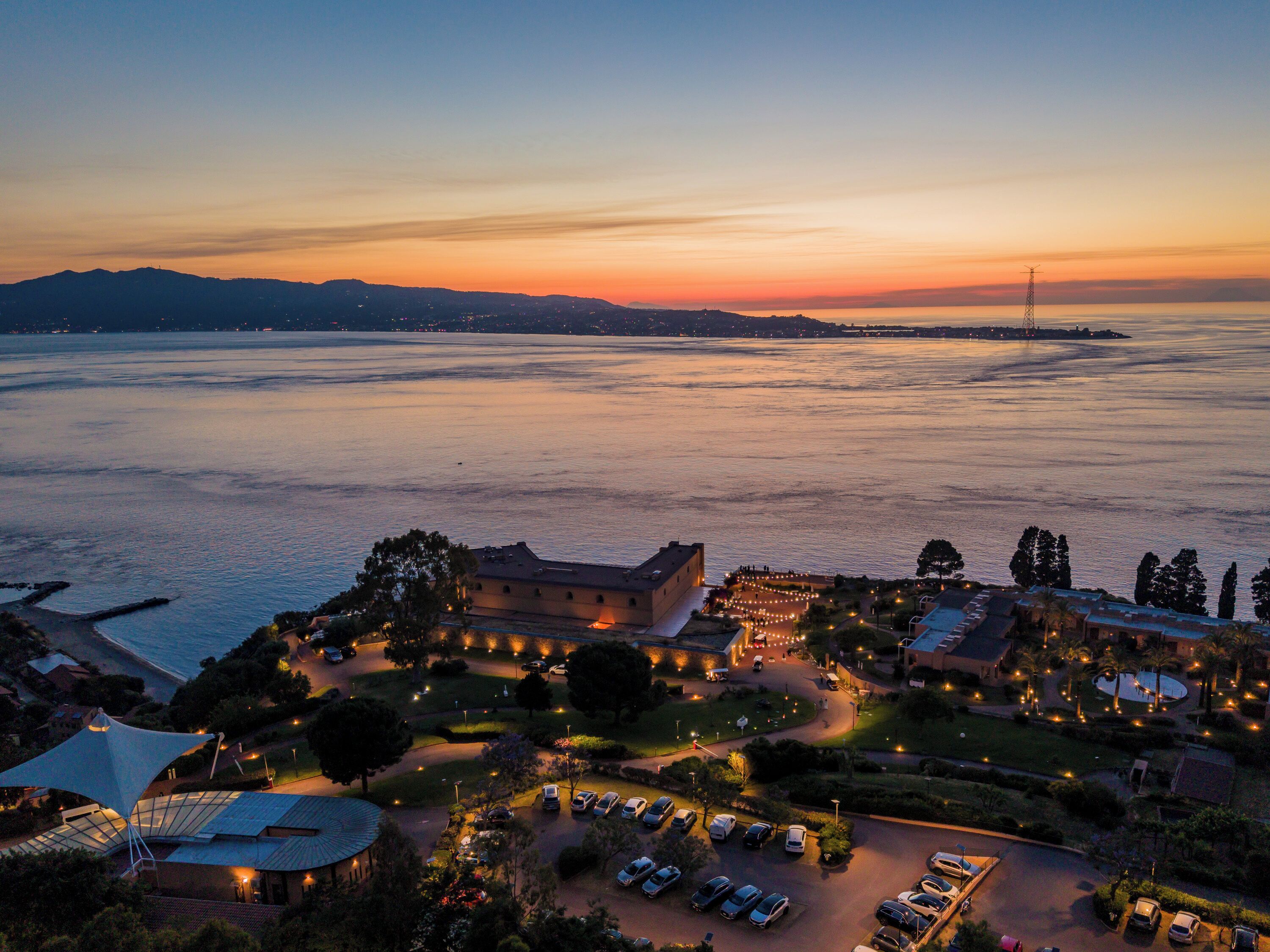 A coastal hotel with a parking area, a large building with a dome, and a view of the sea and distant mountains.