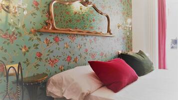 A bedroom with a bed, a chair, a mirror, and a wall with floral wallpaper.