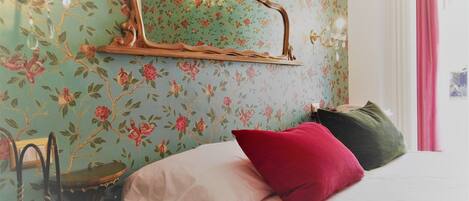 A bedroom with a bed, a chair, a mirror, and a wall with floral wallpaper.