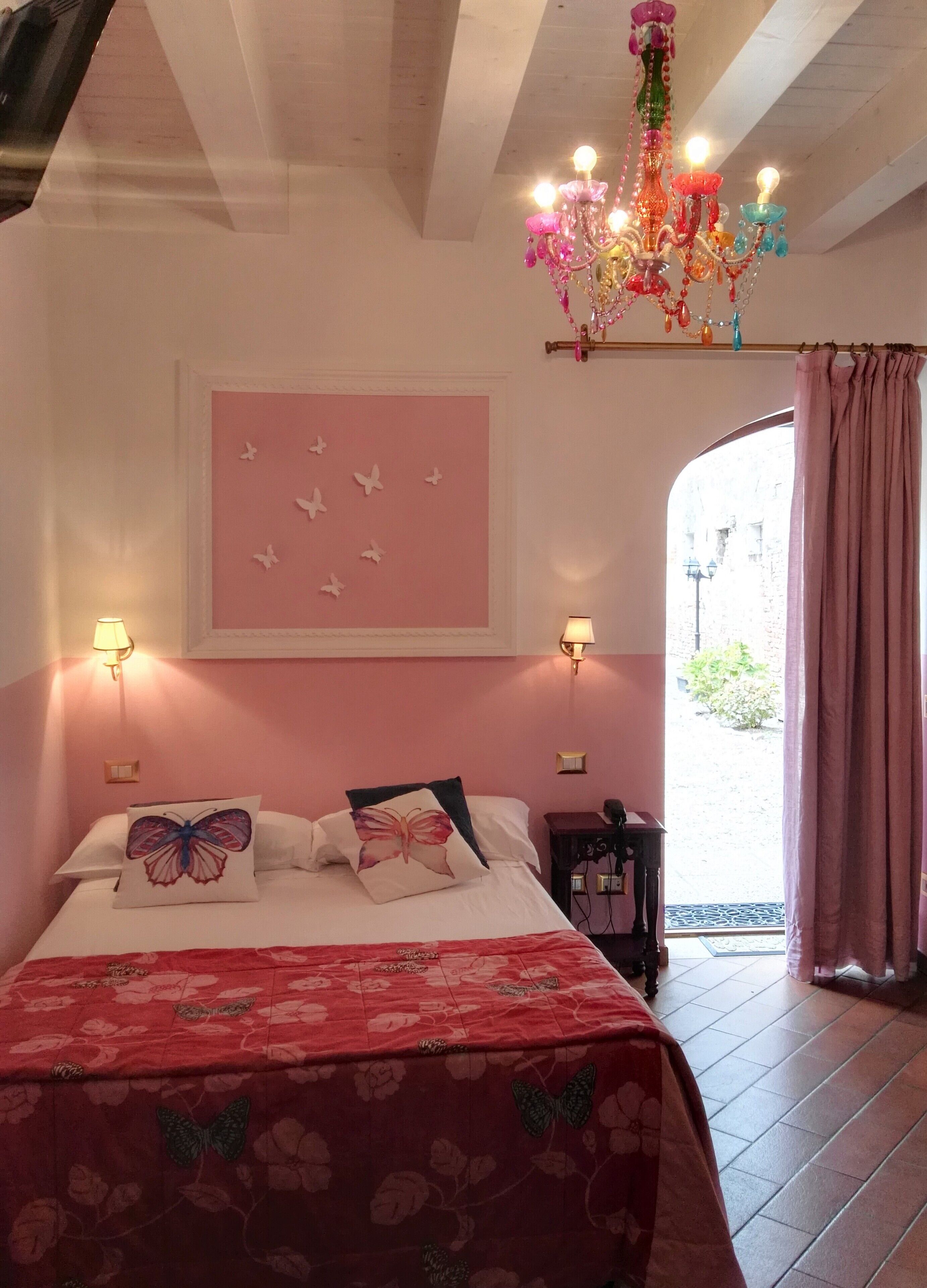 Un dormitorio con cama, almohadas con motivos de mariposas, pared rosa decorada con mariposas, lámpara de araña y ventana con cortinas.