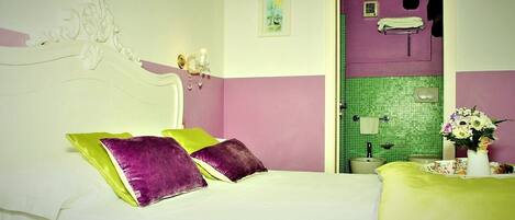 Ein Schlafzimmer mit Bett, Kissen, einem Nachttisch, einer Vase mit Blumen und einem durchgegangenen Tür erkennbaren Badezimmer.