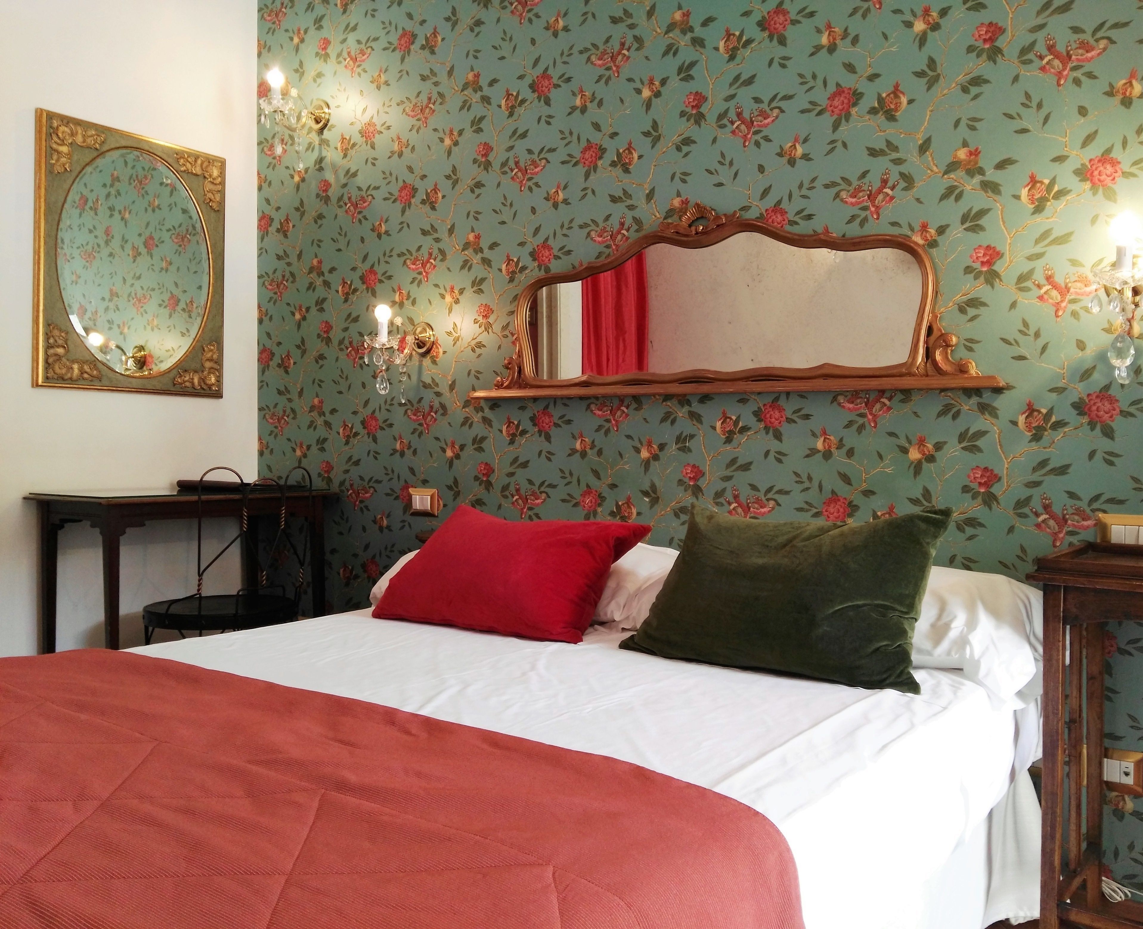 A bed with red and green pillows, a mirror with a gold frame, and floral wallpaper.