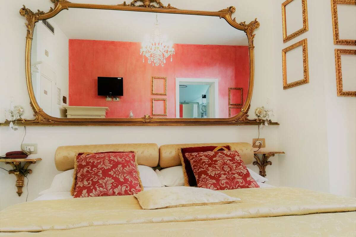 Un dormitorio con un espejo grande y ornamentado, una araña de cristal y una cama con ropa de cama roja y blanca.