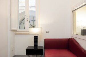 A corner room with a red sofa, a black side table with a lamp, a window with a view of a building, and a wall-mounted light switch.