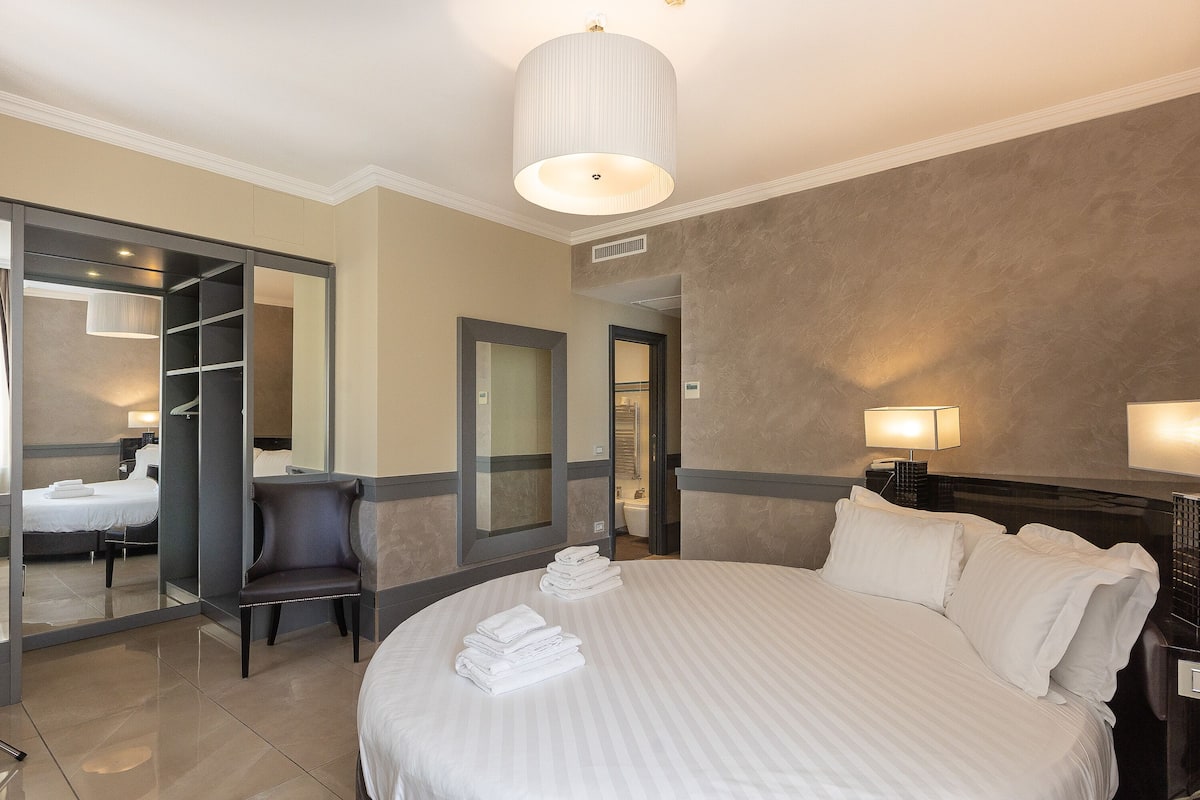 A hotel room with a large bed, a chair, a desk, a mirror, and a bathroom visible through an open door.