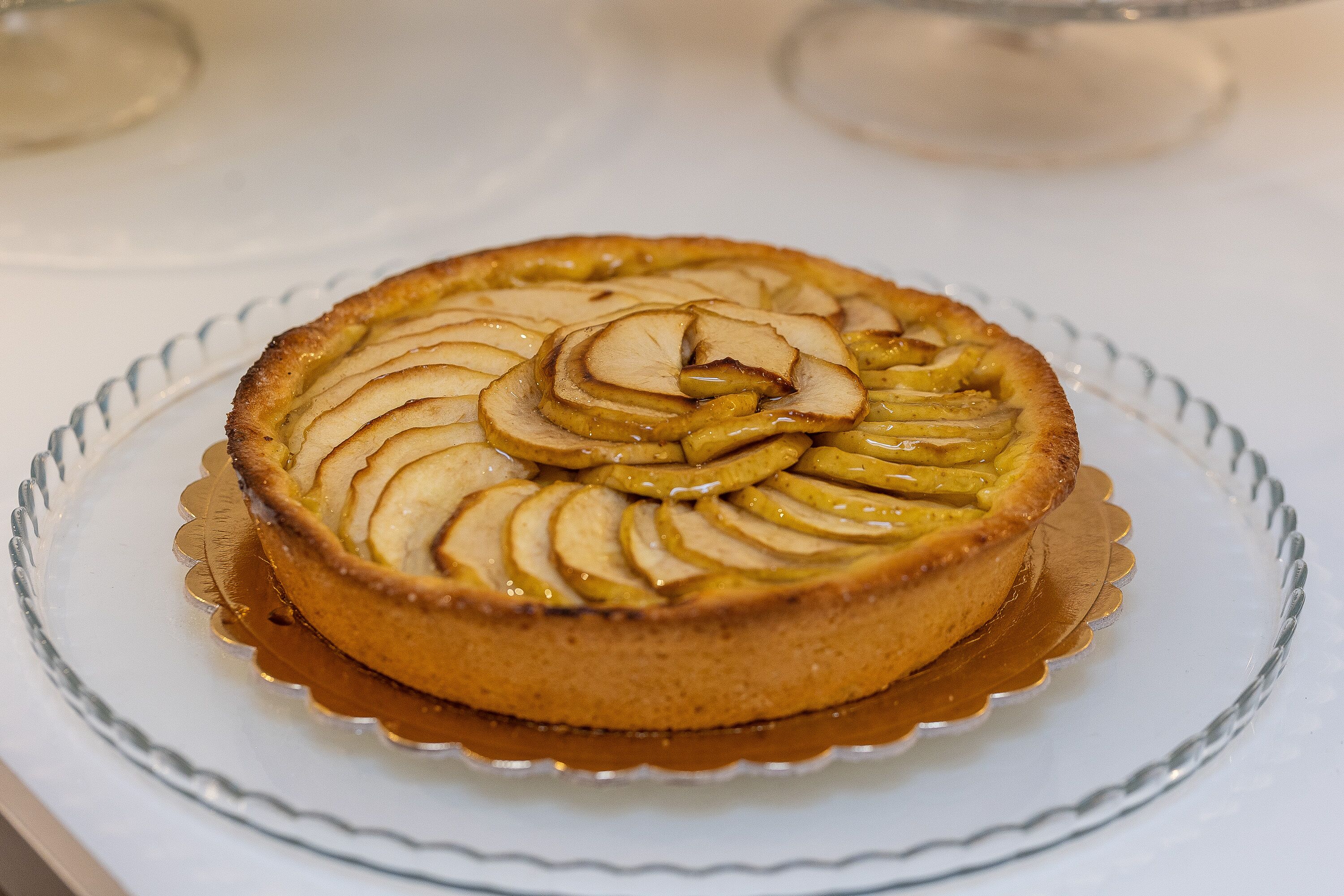 Un pastel redondo con rebanadas de manzana por encima, sobre un plato de vidrio transparente.