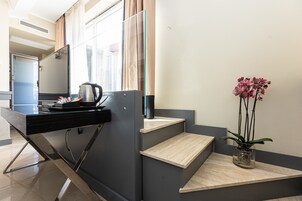 Et moderne hotelværelse med et glas skrivebord, kaffemaskine, en orkidé i potte og udsigt over byen gennem store vinduer.