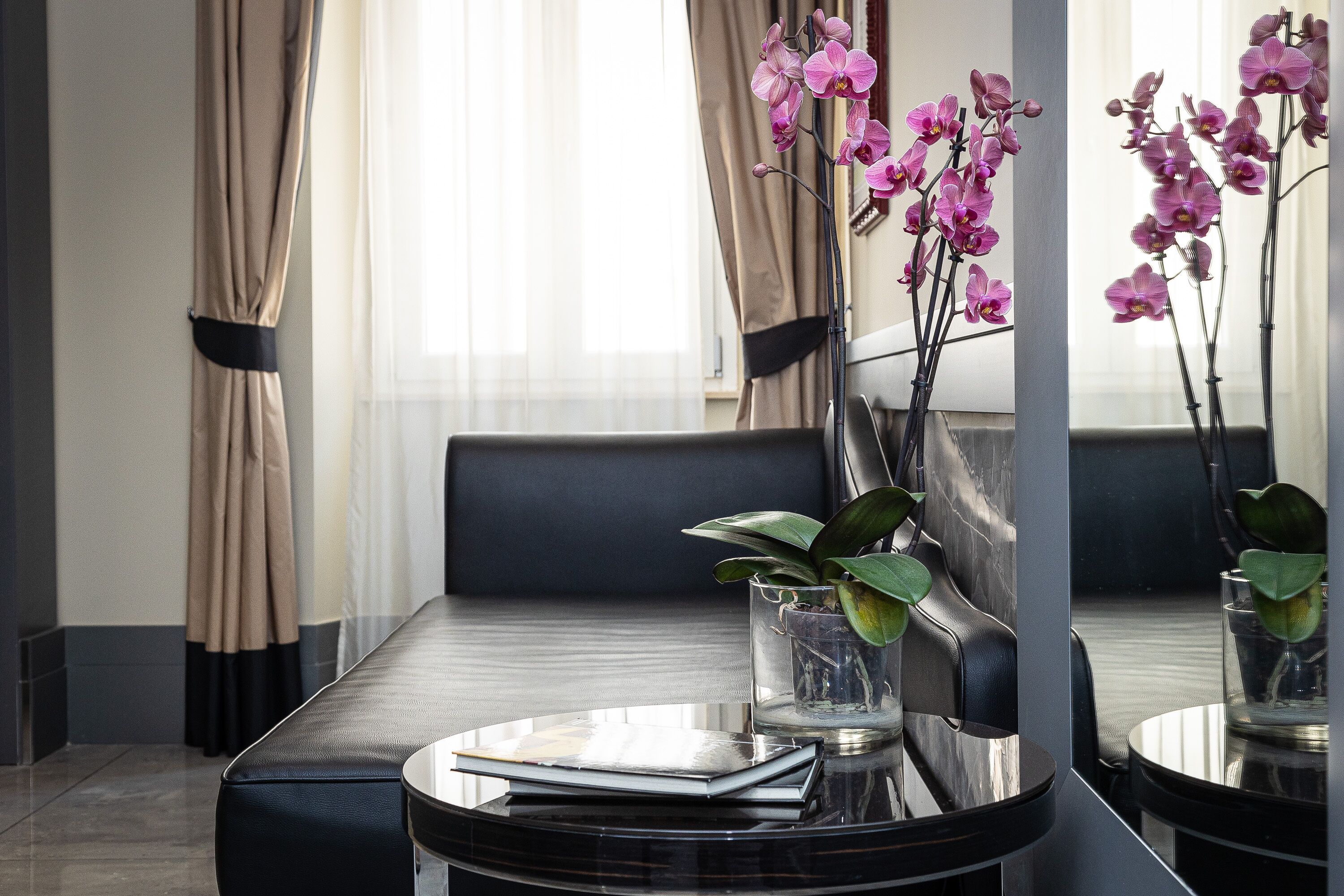 A modern hotel room with a dining area featuring a glass table, a sofa, and a large mirror reflecting the room.