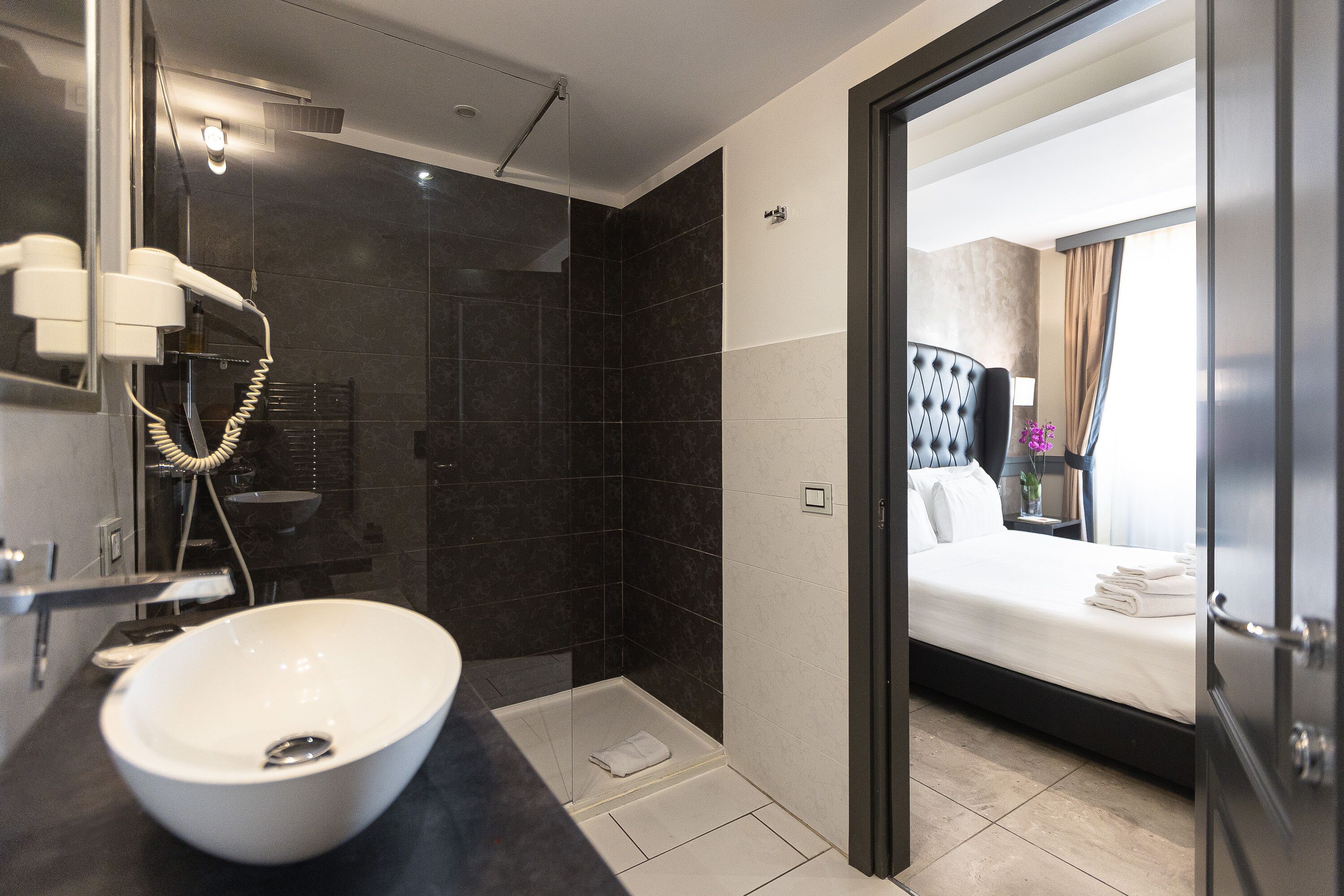 Un baño moderno con un lavamanos grande de color blanco, una ducha con mampara de vidrio y una habitación visible con cama y cortinas.