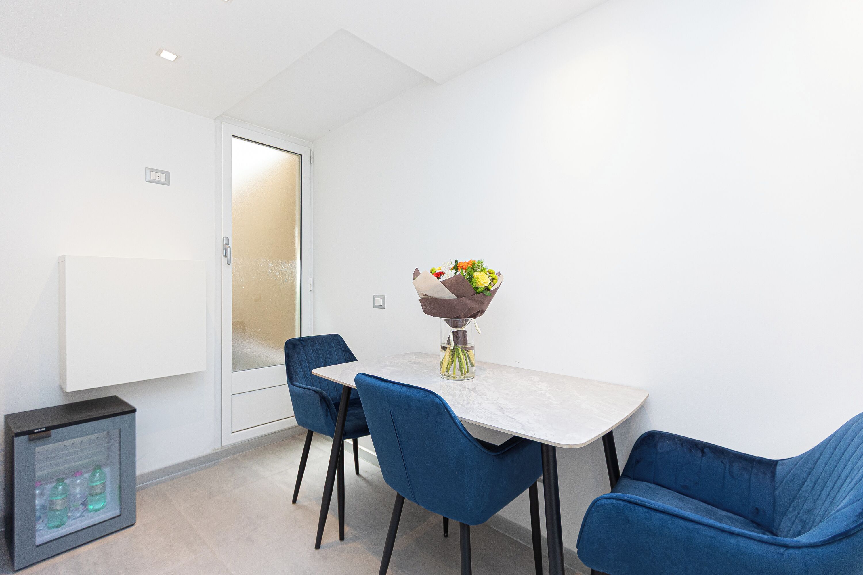 A modern dining area with a white table, two blue chairs, and a vase of flowers.