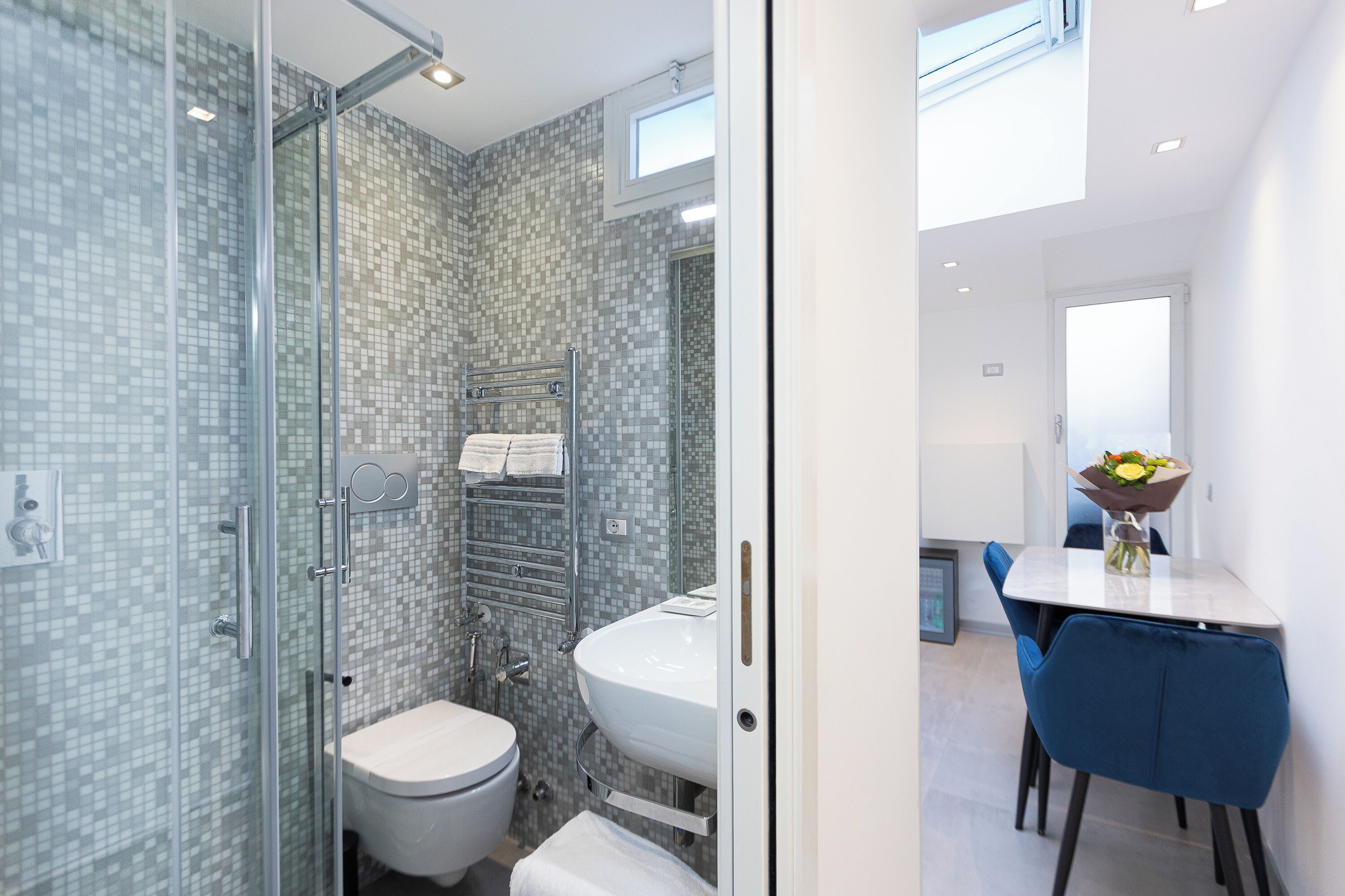 A modern bathroom with a glass shower enclosure, a white toilet, and a sink with a towel rack. There is a dining area with a table and chairs visible through the bathroom door.