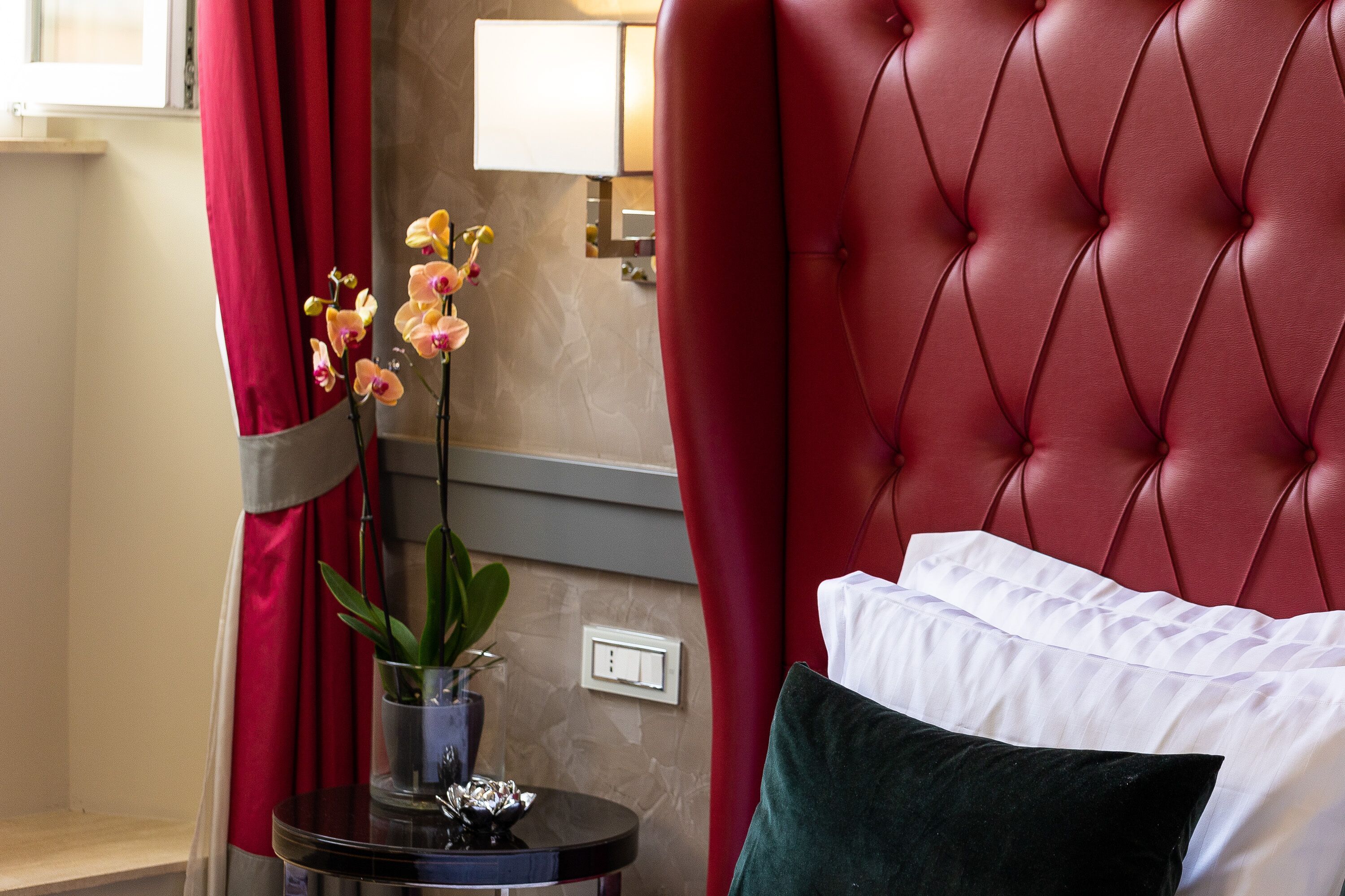 A hotel room with a tufted red headboard, a white pillow, a green pillow, and a vase with orchids on a round table.