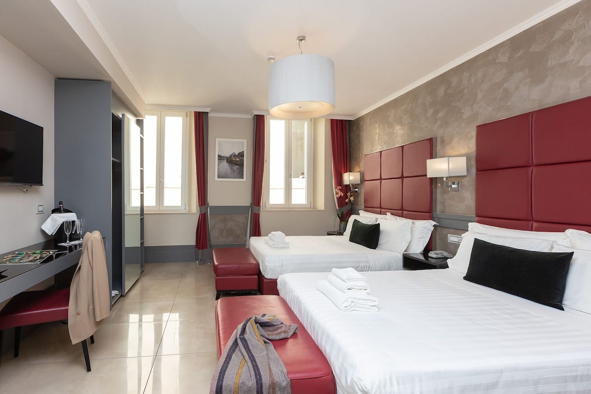 A hotel room with two beds, a desk with a chair, a TV, and a large window with curtains.