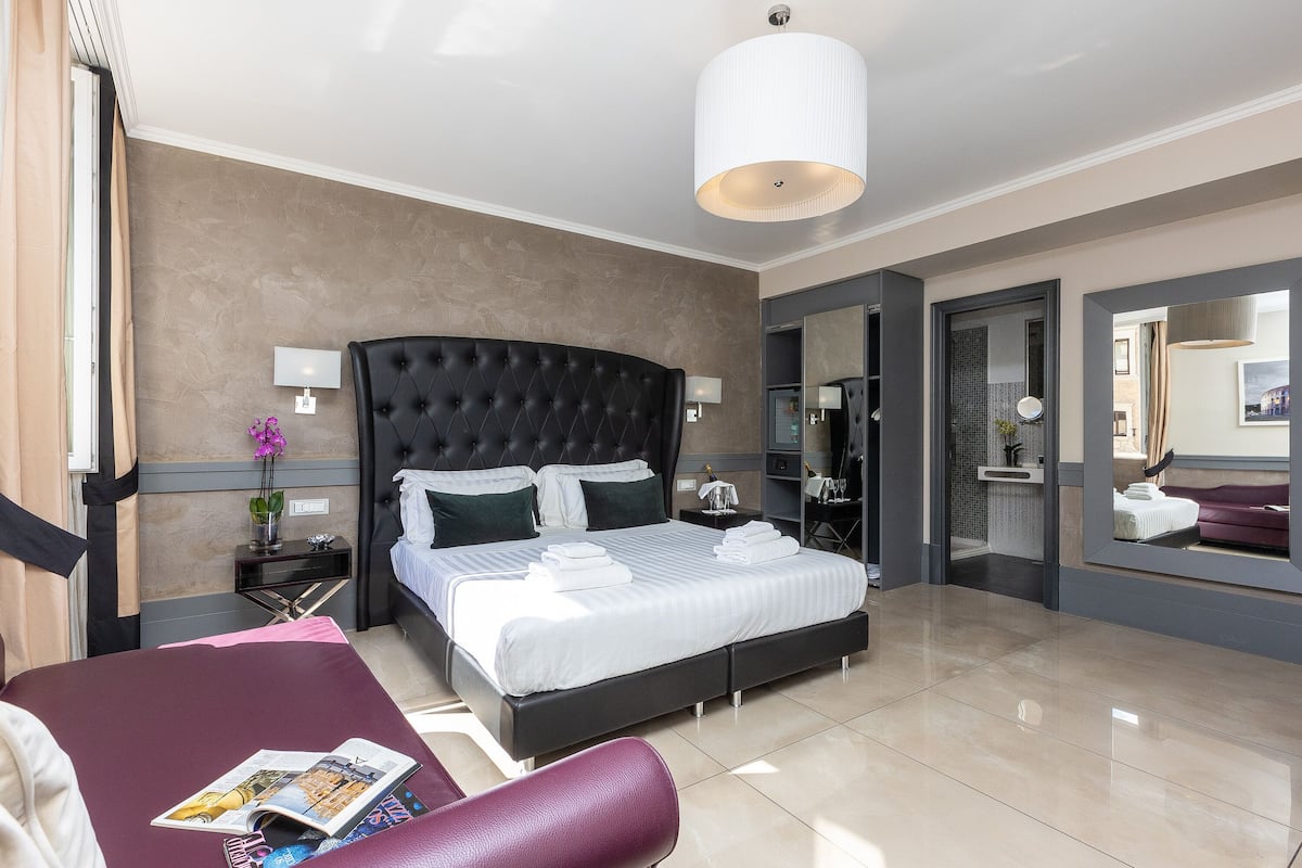 A modern hotel room with a large bed, a bedside table, a chair, and a large mirror.