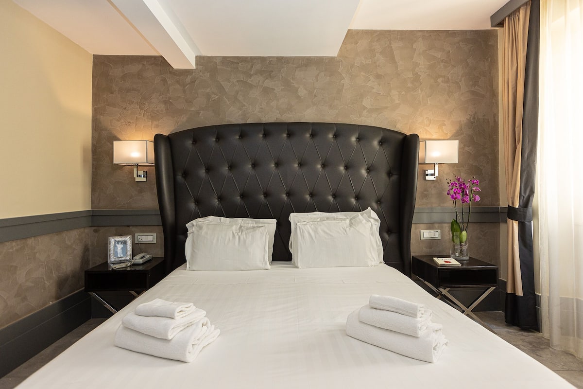 A hotel room with a large bed, two bedside tables, a nightstand, and a vase with flowers.