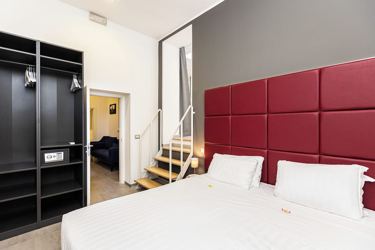 A modern hotel room with a large bed, a red headboard, a wardrobe, and a staircase leading to another room.