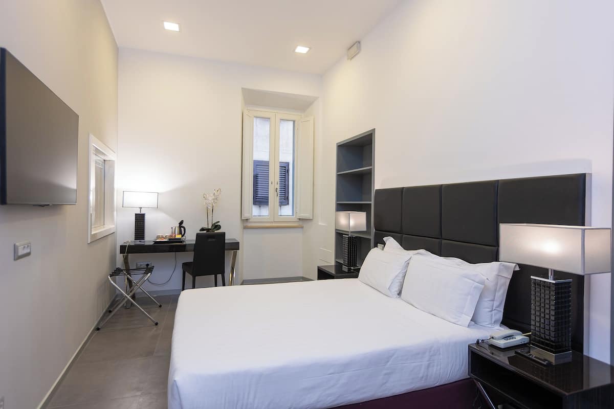 A hotel room with a bed, a desk with a chair, a TV, a window, and a nightstand with a lamp.