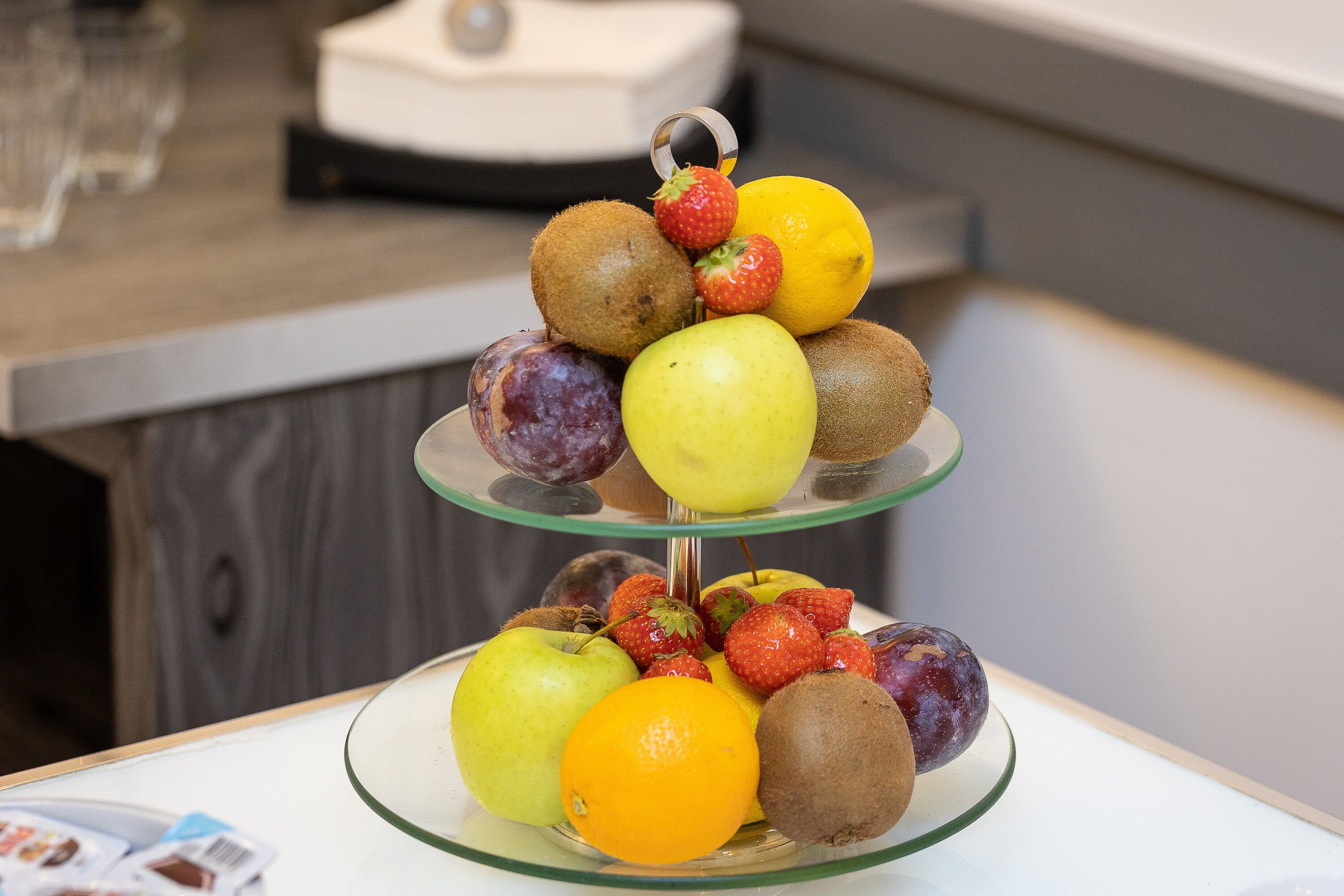 A three-tiered fruit stand with apples, kiwis, strawberries, and an orange on a glass table.