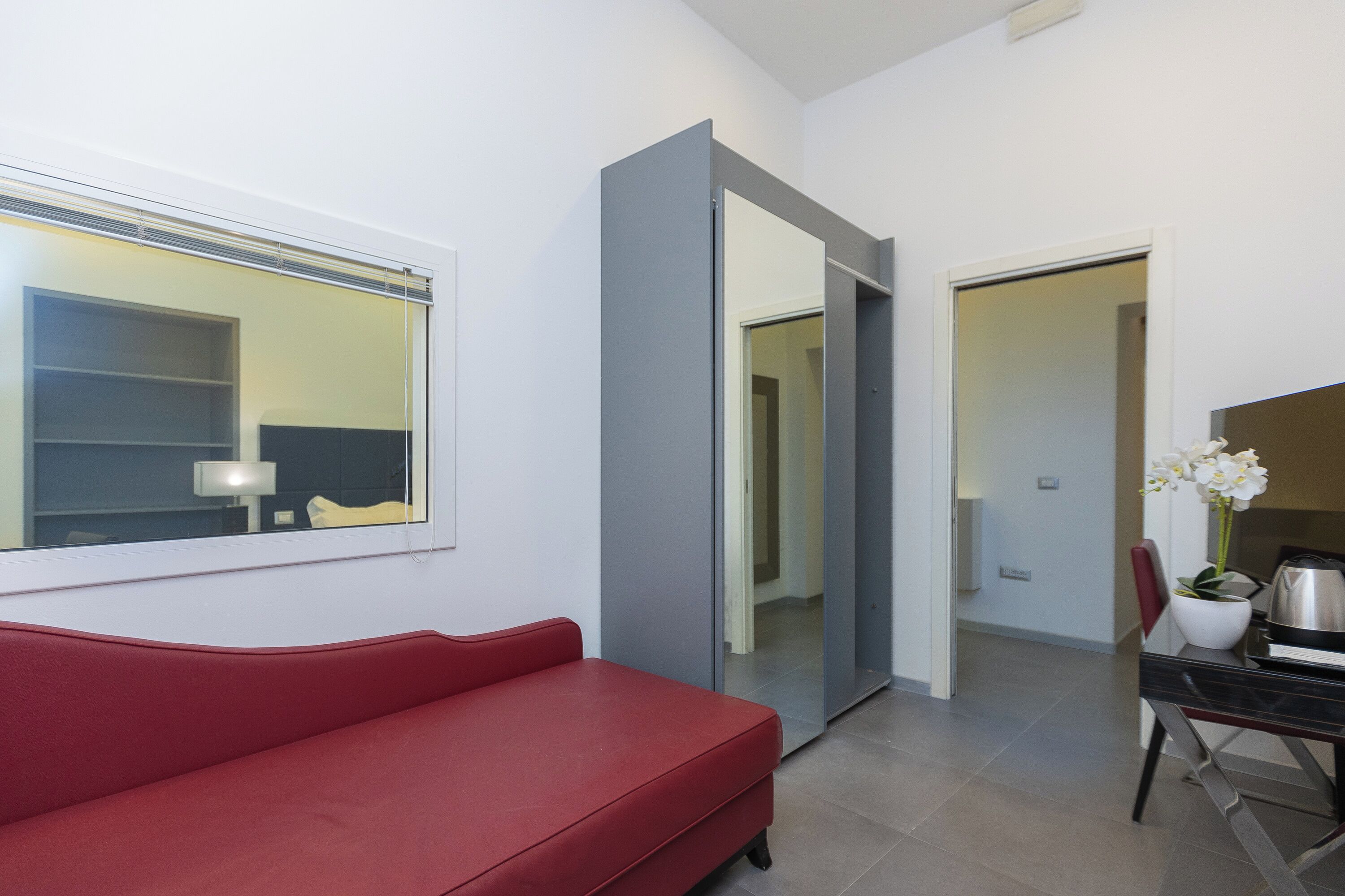 Una habitación de hotel moderna con un sofá rojo, un espejo grande, un escritorio con cafetera y una orquídea en maceta.