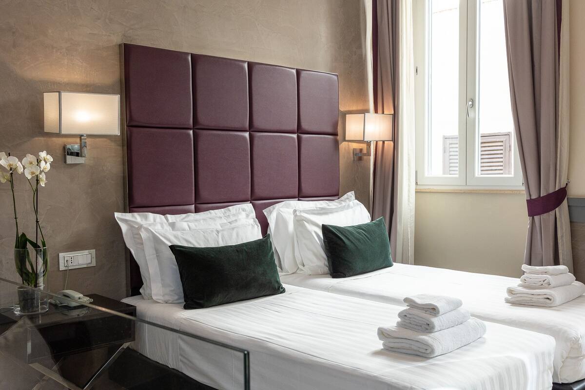 A hotel room with a large padded headboard, two beds with white linens and green pillows, a bedside table with a lamp, and a window with curtains.