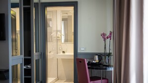 A hotel room with a glass door leading to a bathroom, a desk with a chair, and a vase with flowers. - Hotel Castellino Roma (Rome)