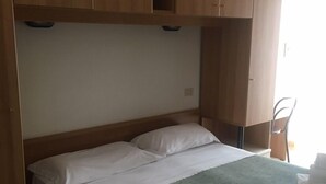 A single bed with a green bedspread and white linens, wooden wardrobe above, and a chair by the window.