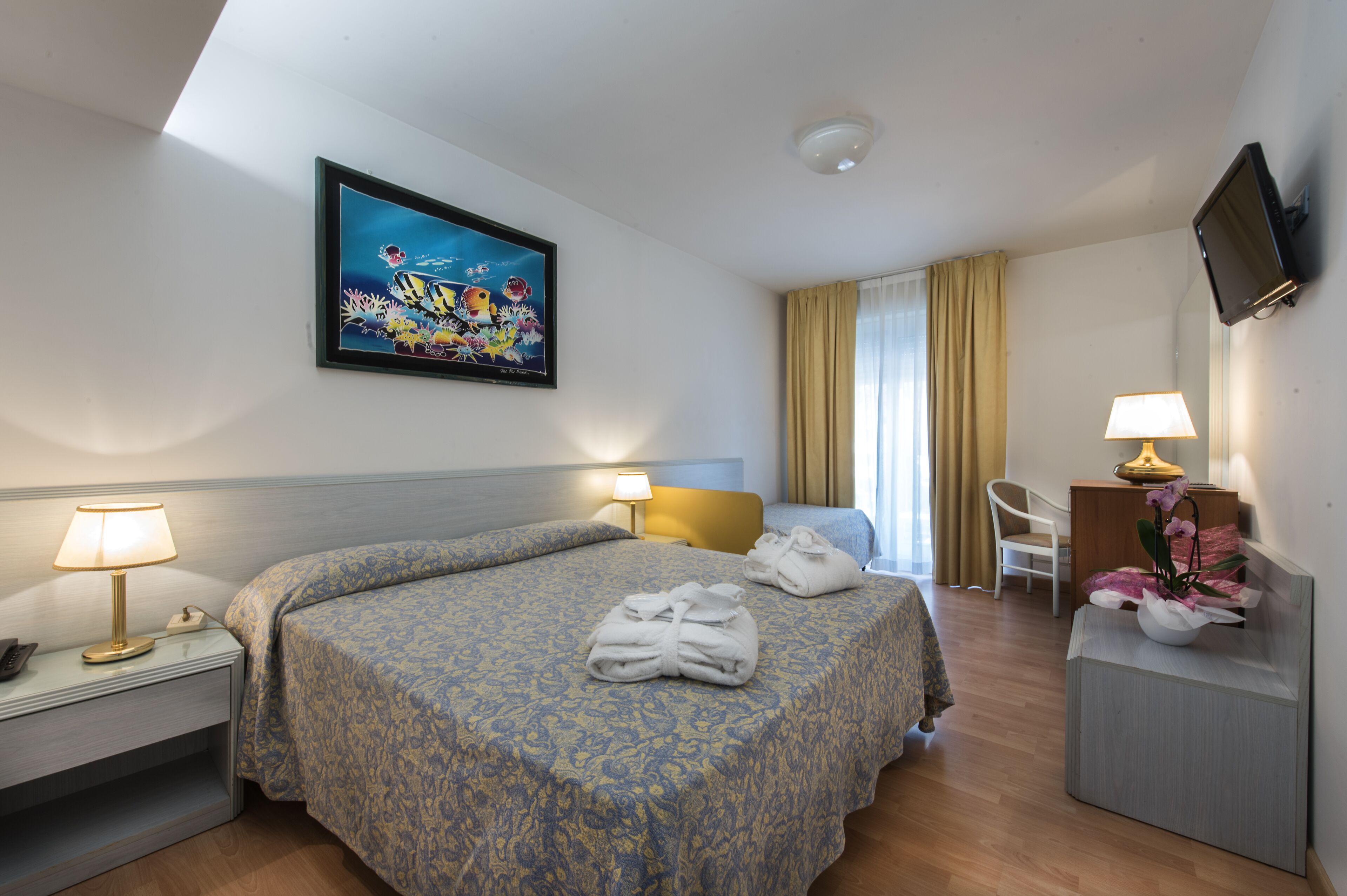 Une chambre d’hôtel comprenant un lit, deux lampes de chevet, un téléviseur à écran plat fixé au mur, un bureau avec une lampe, une chaise et un tableau représentant un paysage tropical.