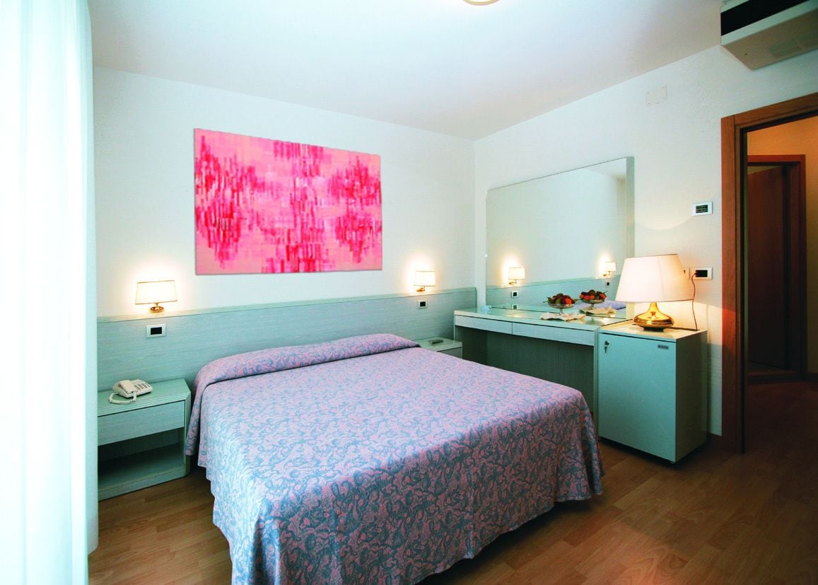 Une chambre d’hôtel avec un lit, des tables de chevet, un bureau, un miroir et un tableau abstrait rose accroché au mur.