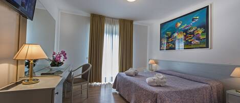 A hotel room with a bed, a desk with a lamp, a chair, a painting on the wall, and a window with curtains.